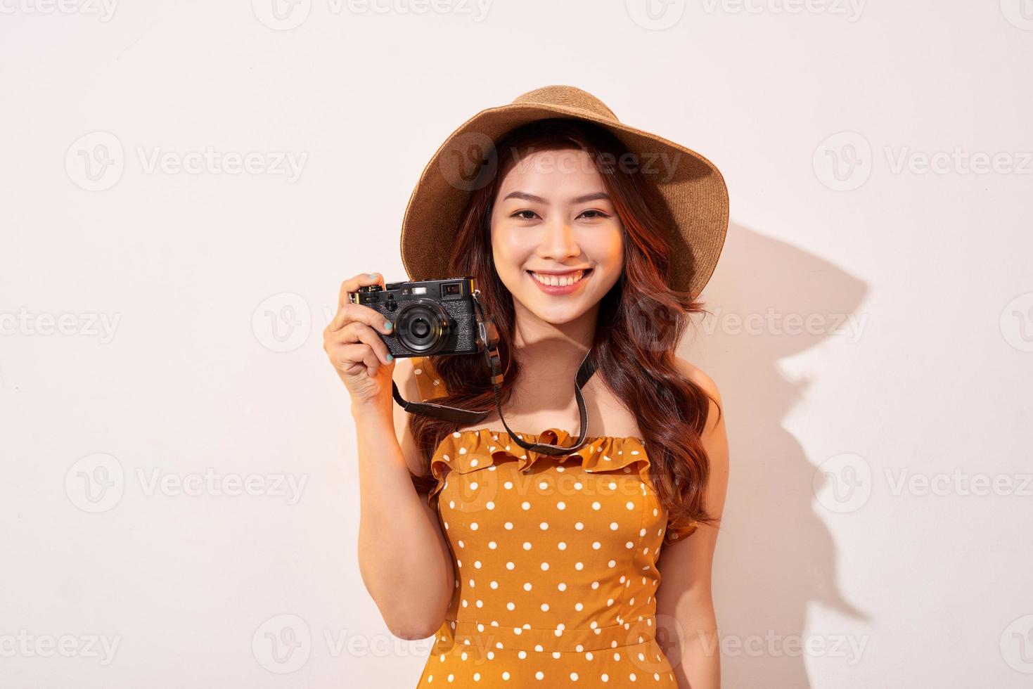 Portrait of cheerful smiling young woman taking photo with inspiration and wearing summer dress. Girl holding retro camera. Model posing on beige background in hat