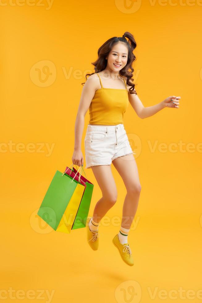 Joyful Teen Summer Girl Carrying Shopping Bags And Jumping In Air Over orange Background, Full-Length Shot With Copy Space photo