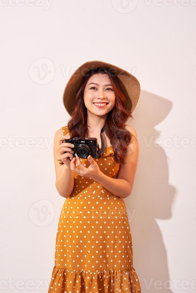 Portrait of cheerful smiling young woman taking photo with inspiration and wearing summer dress. Girl holding retro camera. Model posing on beige background in hat