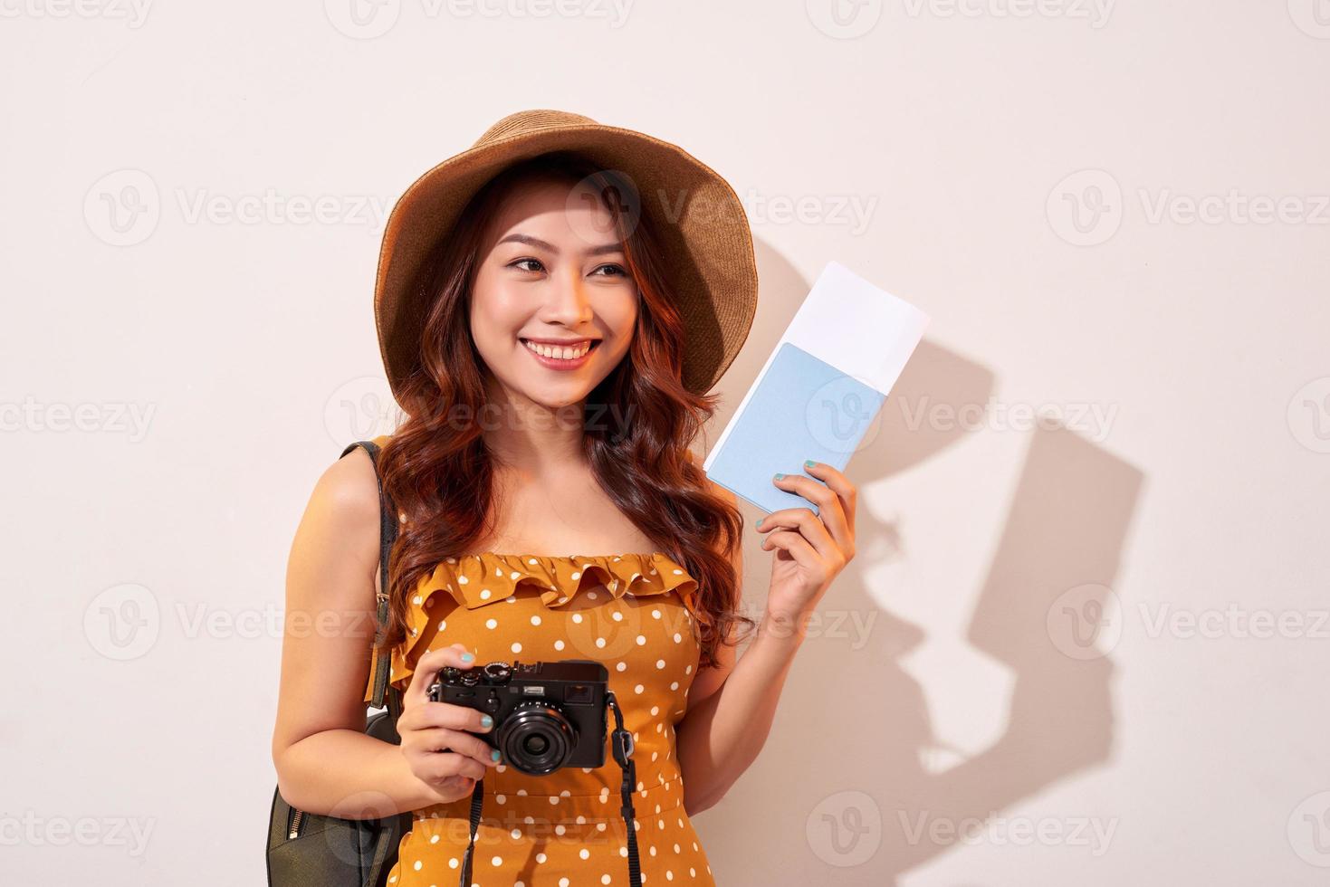 retrato de una joven feliz con sombrero sosteniendo una cámara y mostrando el pasaporte mientras se encuentra aislada sobre un fondo beige foto