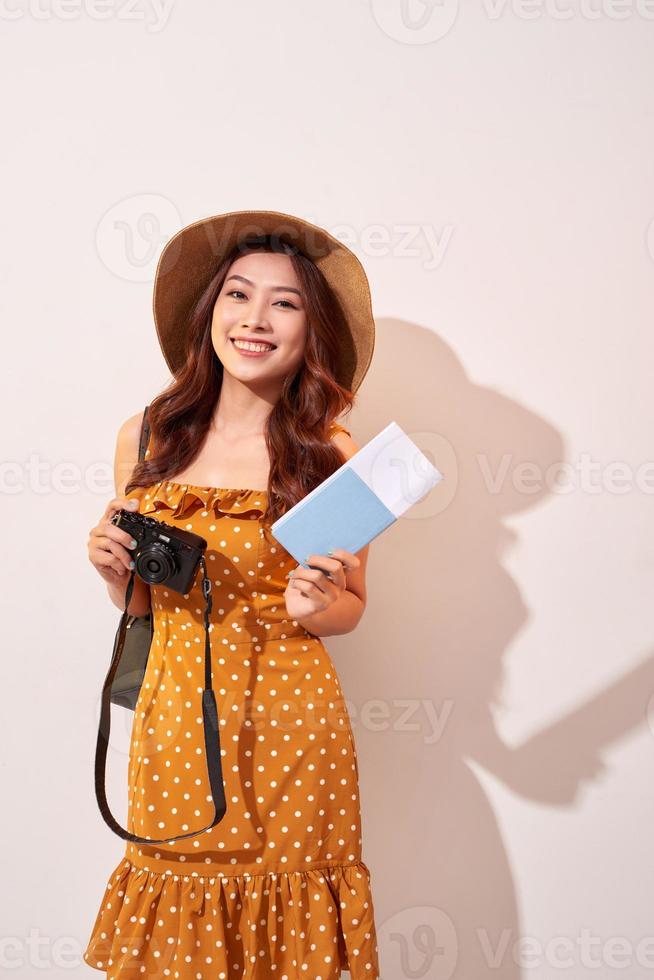 retrato de una joven feliz con sombrero sosteniendo una cámara y mostrando el pasaporte mientras se encuentra aislada sobre un fondo beige foto