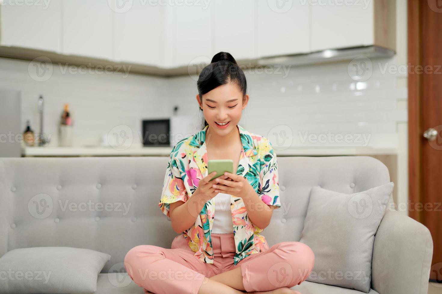 Smiling teenager girl relax sit on sofa at home using cellphone texting chatting with friend photo