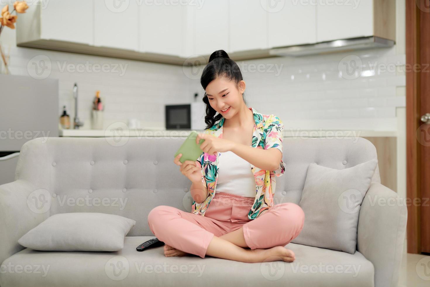 Smiling young asian woman using mobile phone app playing game, relax on sofa. photo