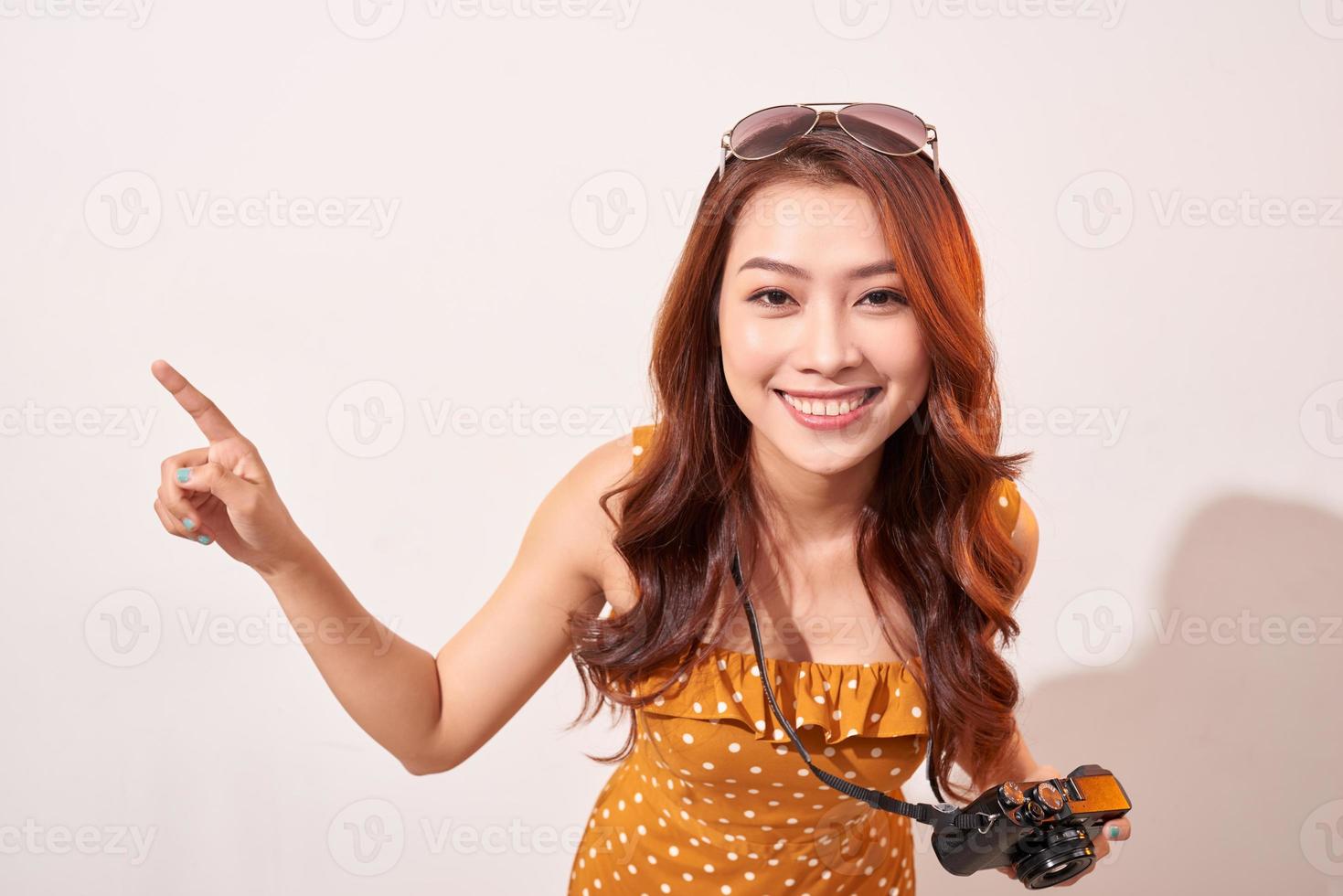 Tourist standing isolated on beige background and joyfully pointing to the sky. Lady using camera while travel. Independent travel in Asia concept. photo