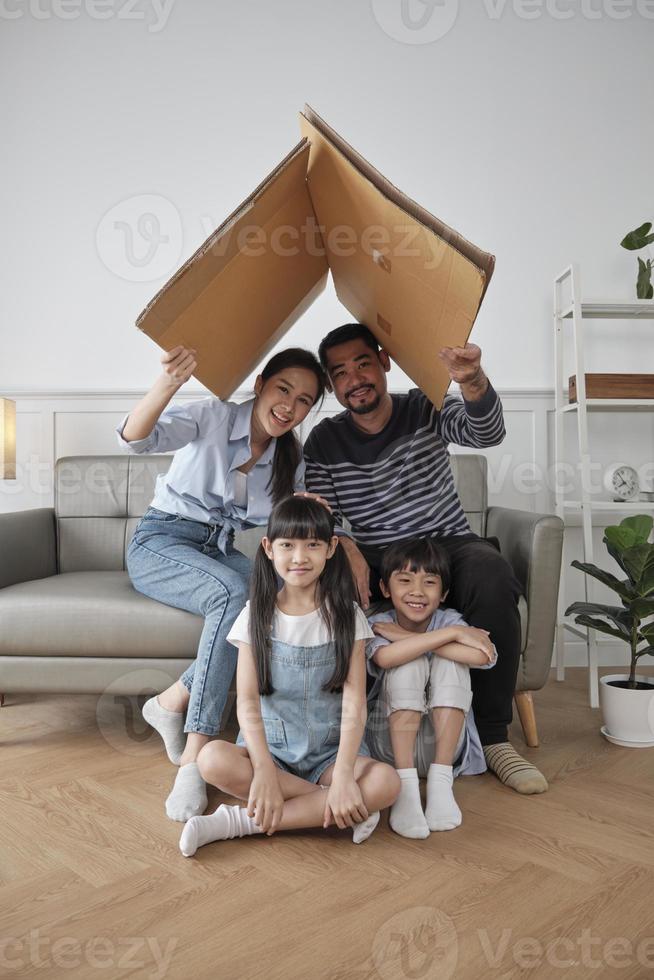 Asian Thai family, adult dad, mum, and little children happily fun playing in a white living room, built cardboard paper safety house with imagination, lovely weekend and wellbeing domestic lifestyle. photo