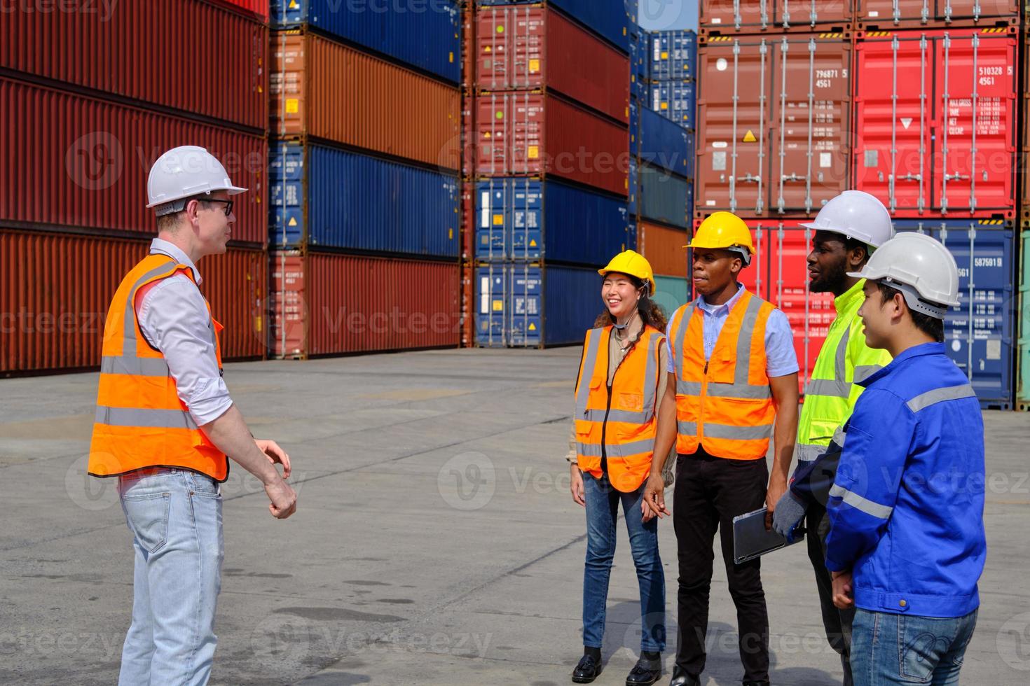 A group of multiracial workers team in safety uniforms discuss with Caucasian manager at logistics dock with many stacks of containers, shipping goods, import and export cargo transportation industry. photo