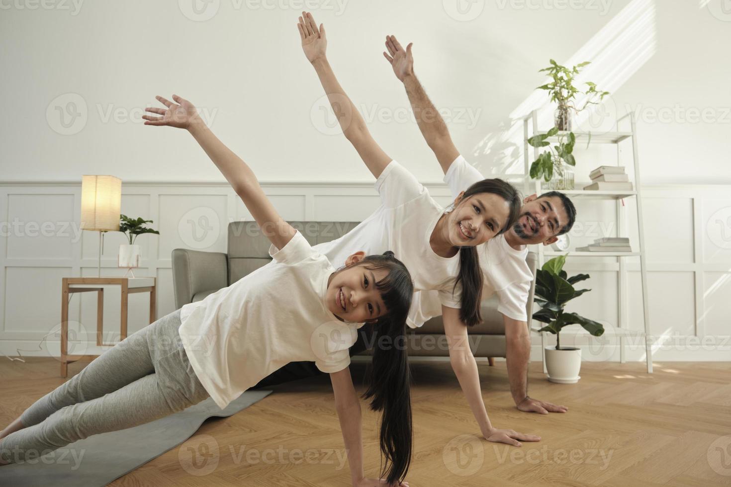 Asian Thai parents and daughter fitness training exercise and practice yoga on living room floor, lovely rowing together for health and wellness, and happy domestic home lifestyle on family weekend. photo