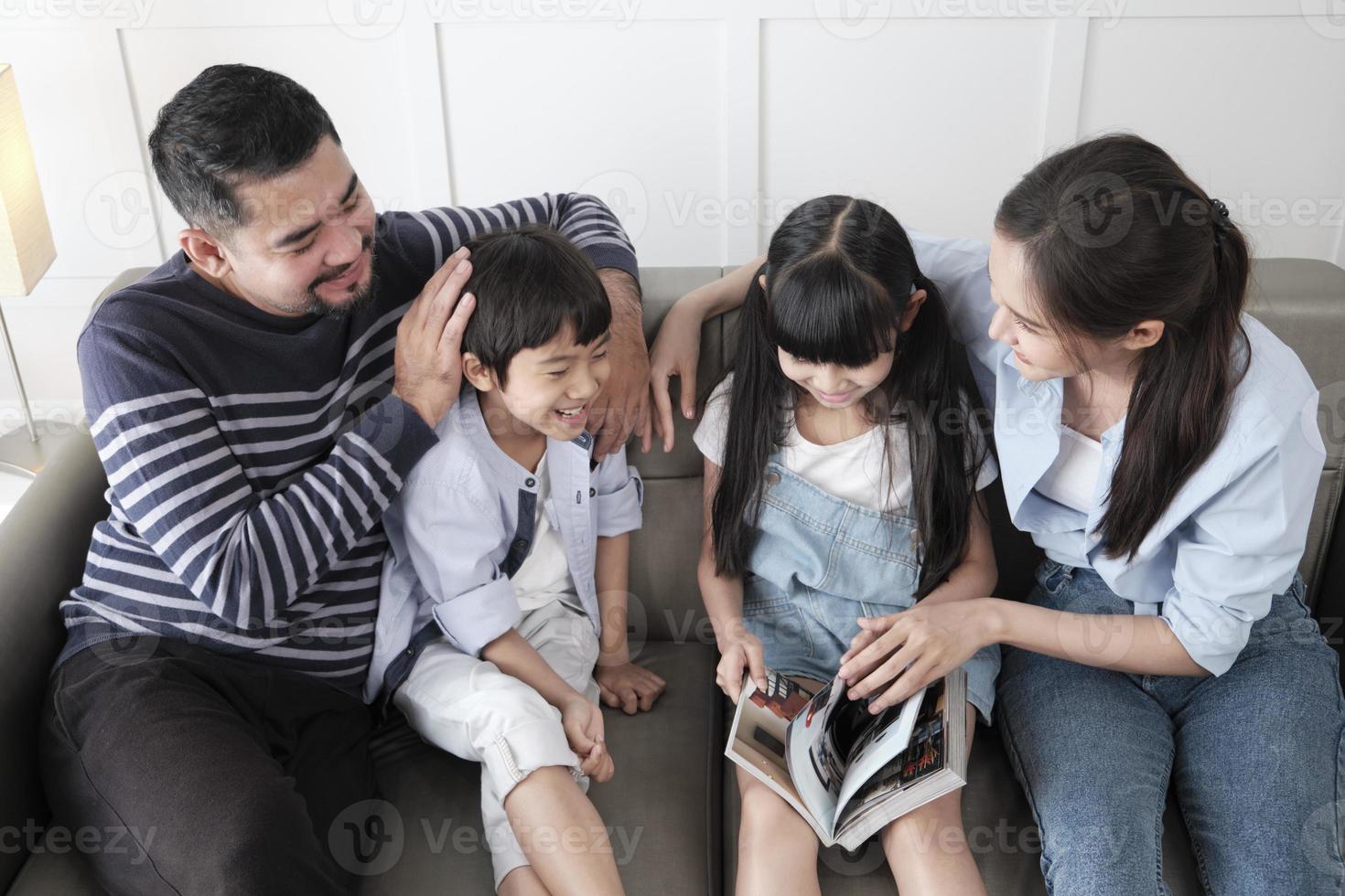 Asian Thai family, adult dad, mum, and children happiness home living relaxing activities and reading book together, leisure on sofa in white room house, lovely weekend, wellbeing domestic lifestyle. photo