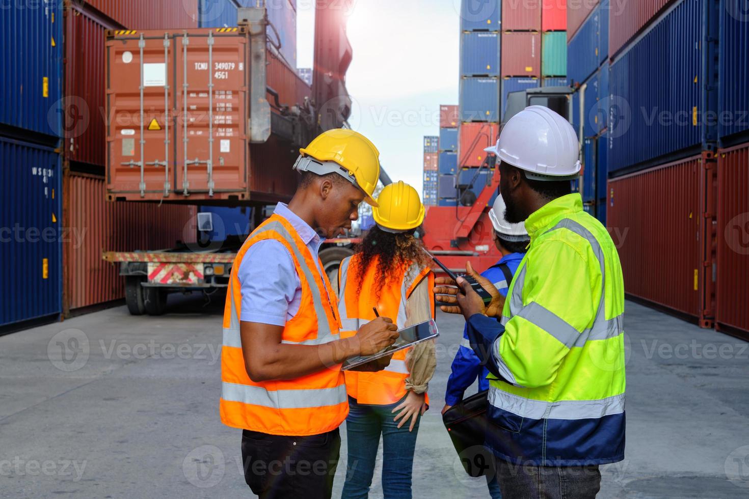 los trabajadores afroamericanos y los equipos con uniformes de seguridad y cascos usan walkie-talkies, trabajan en grúas logísticas con pilas de contenedores, control de carga, envío de mercancías e industria de transporte de carga. foto