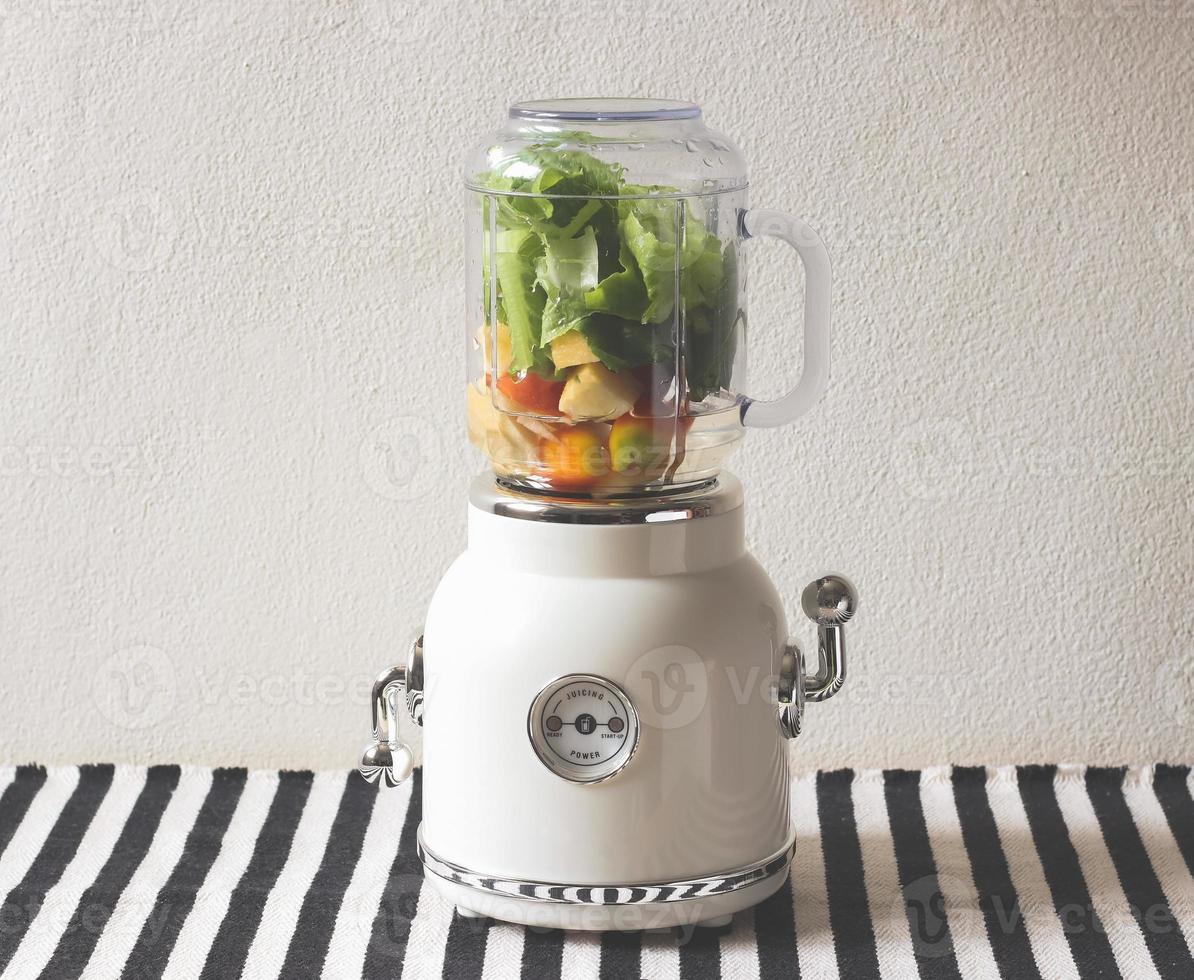 white vintage blender or smoothie maker withe vegetables, tomatos, apples and water   on  black and white stripe table cloth and white wall. photo