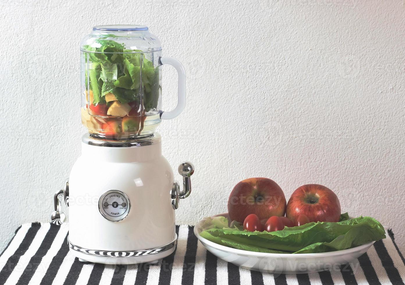 white vintage blender or smoothie maker  with a plate of vegetables, tomatoes and apples on  black and white stripe table cloth and white wall. Healthy drink making. photo