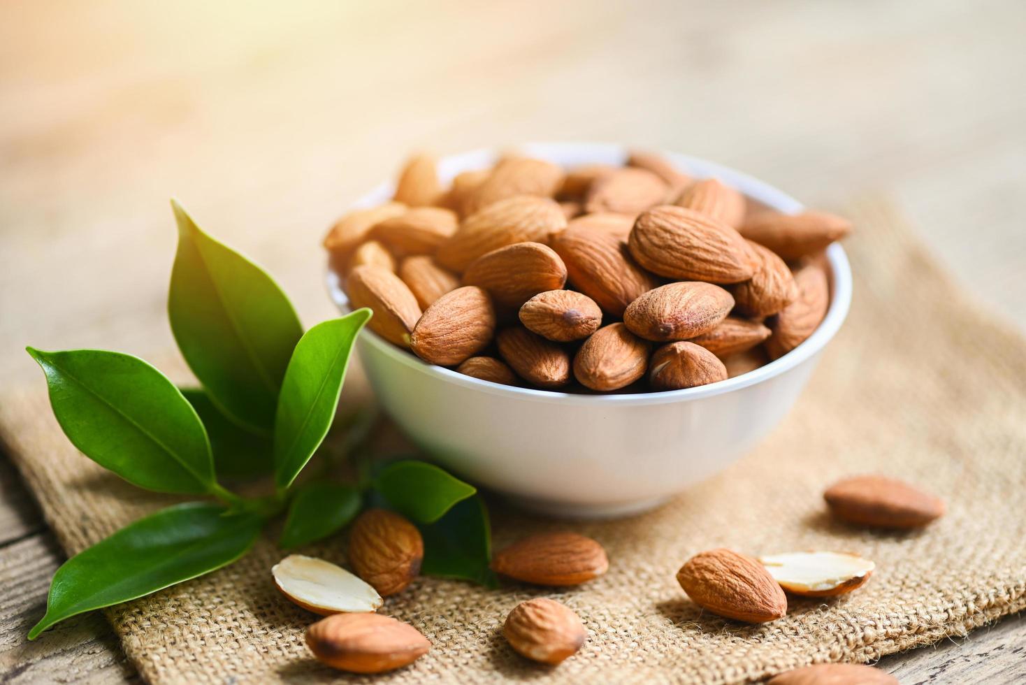 Almonds nuts on white bowl and green leaf on sack background, Delicious sweet almonds on the wooden table , roasted almond nut for healthy food and snack photo