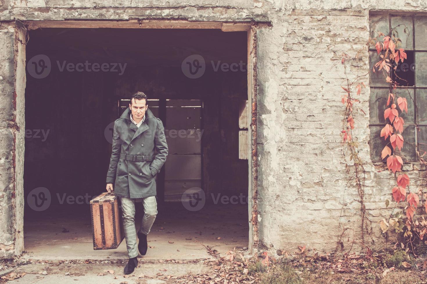 Confident man carrying old leather suitcase and getting out of abandoned building. photo