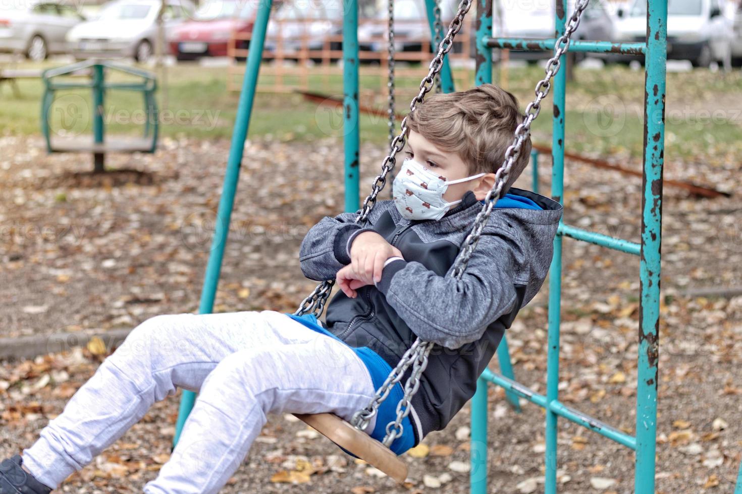 Pensive boy swinging on the playground while wearing face mask due to coronavirus pandemic. photo