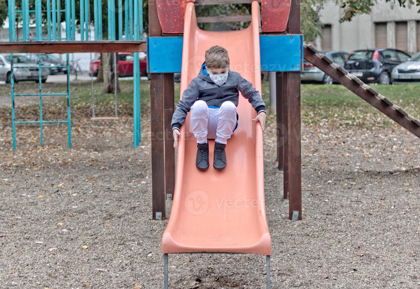Small boy with protective face mask sliding on the playground. photo