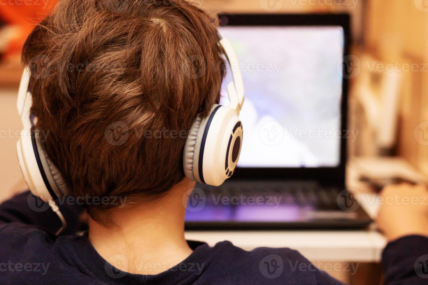 Rear view of kid with headphones following online class over laptop at home. photo