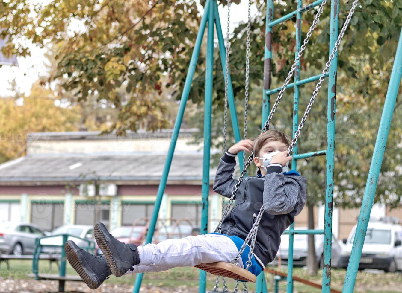 Small boy swinging at the playground and wearing protective face mask. photo