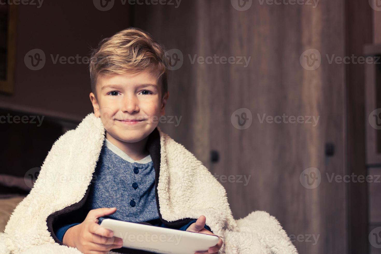 Smiling boy wrapped in blanket using touchpad in bedroom. photo