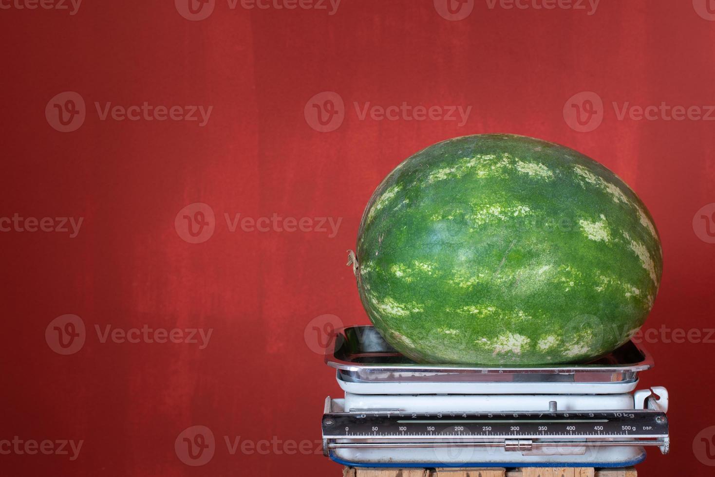 An old kitchen scale stands against a red background with space for text. There is a large green watermelon on the scales photo