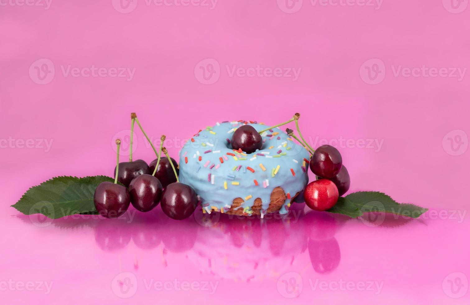 Close-up of a blue donnought with red cherries reflected on a pink background. There are also cherry leaves to see. There is room for text photo