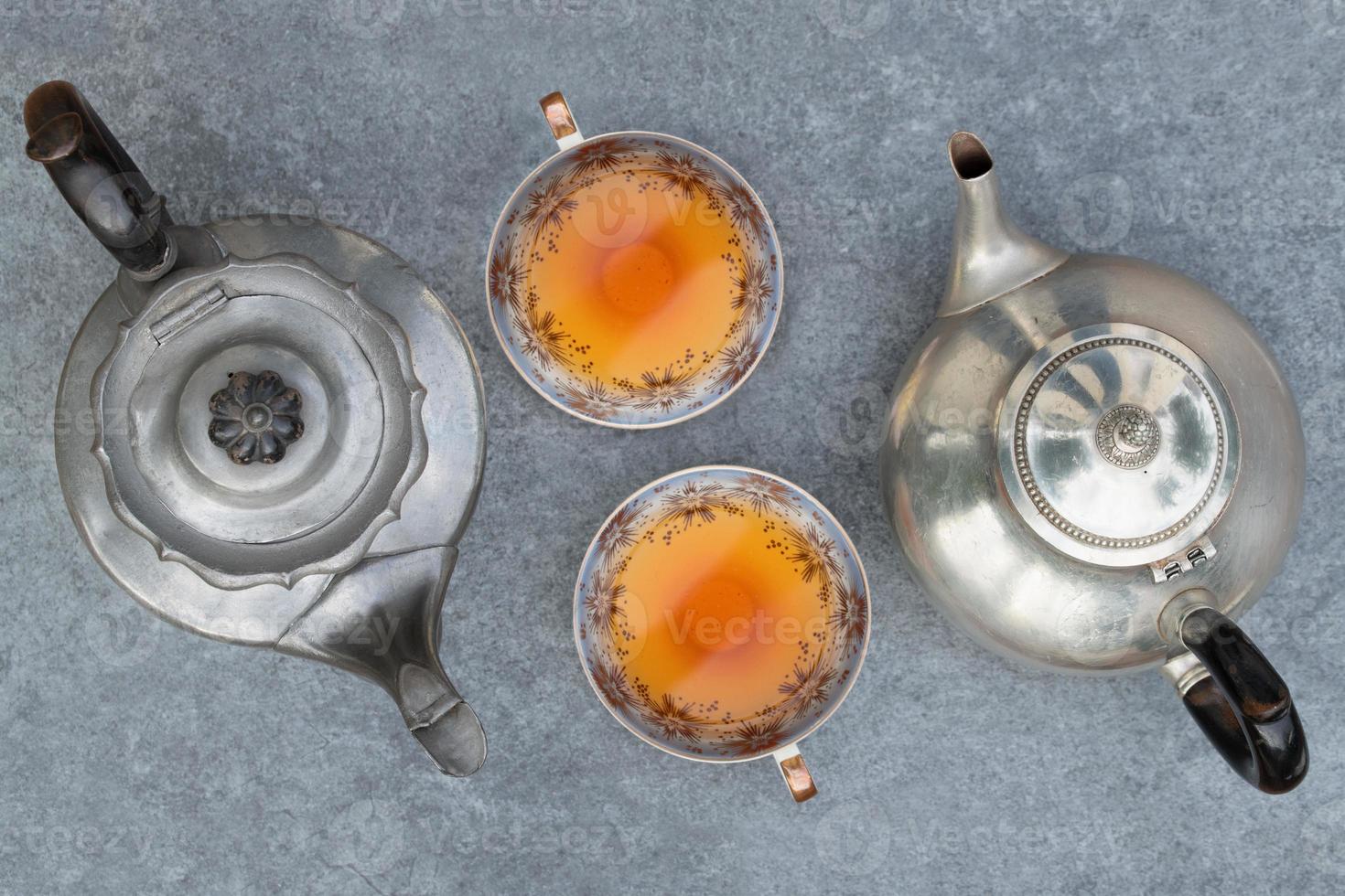 Two different old metal teapots stand on a gray background. Two identical ceramic teacups are filled with tea and stand next to each other. Tea for two photographed from above. photo