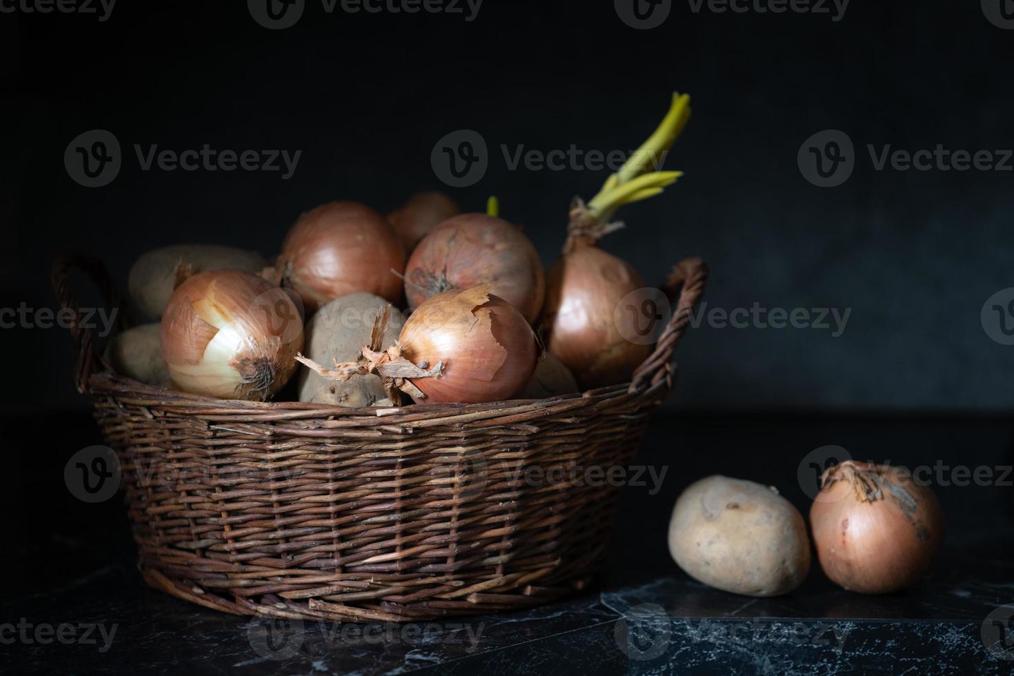 There is a small wicker basket in a kitchen. In the basket are potatoes and onions. The underground and the background are gray and dark. photo