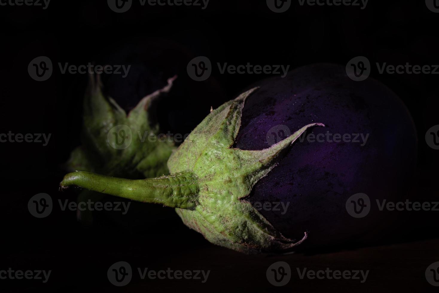 Dark shot of two purple aubergines with green stems lying side by side against a dark background. photo