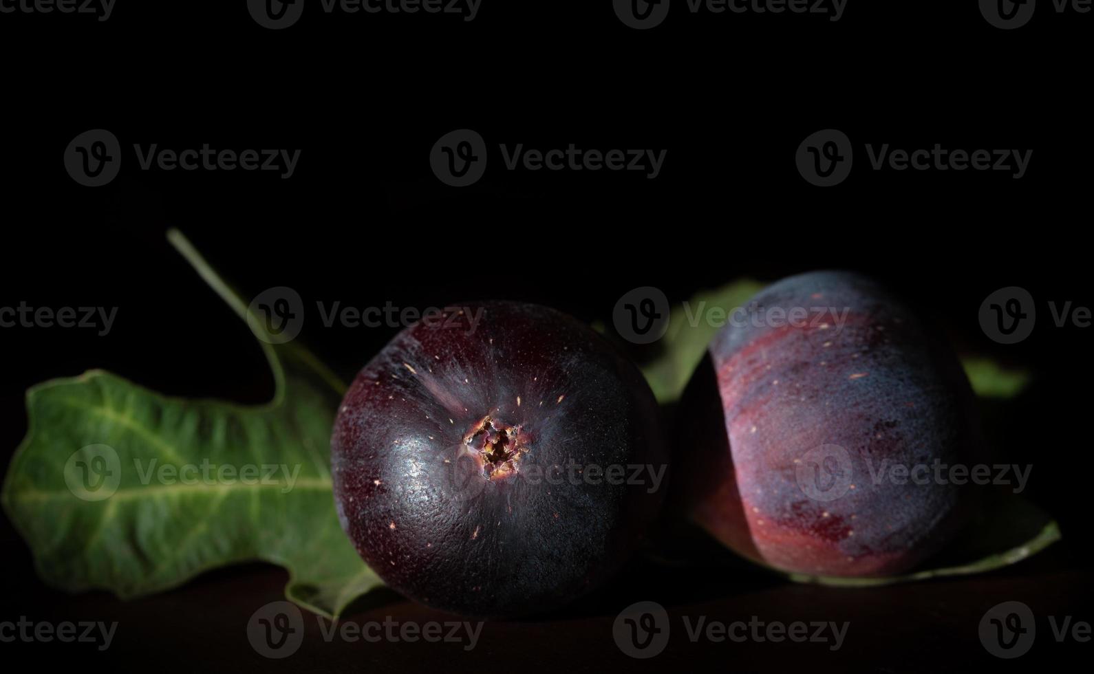 Dark shot of two red figs lying side by side on a table. A few fig leaves in the background. The room is dark. photo