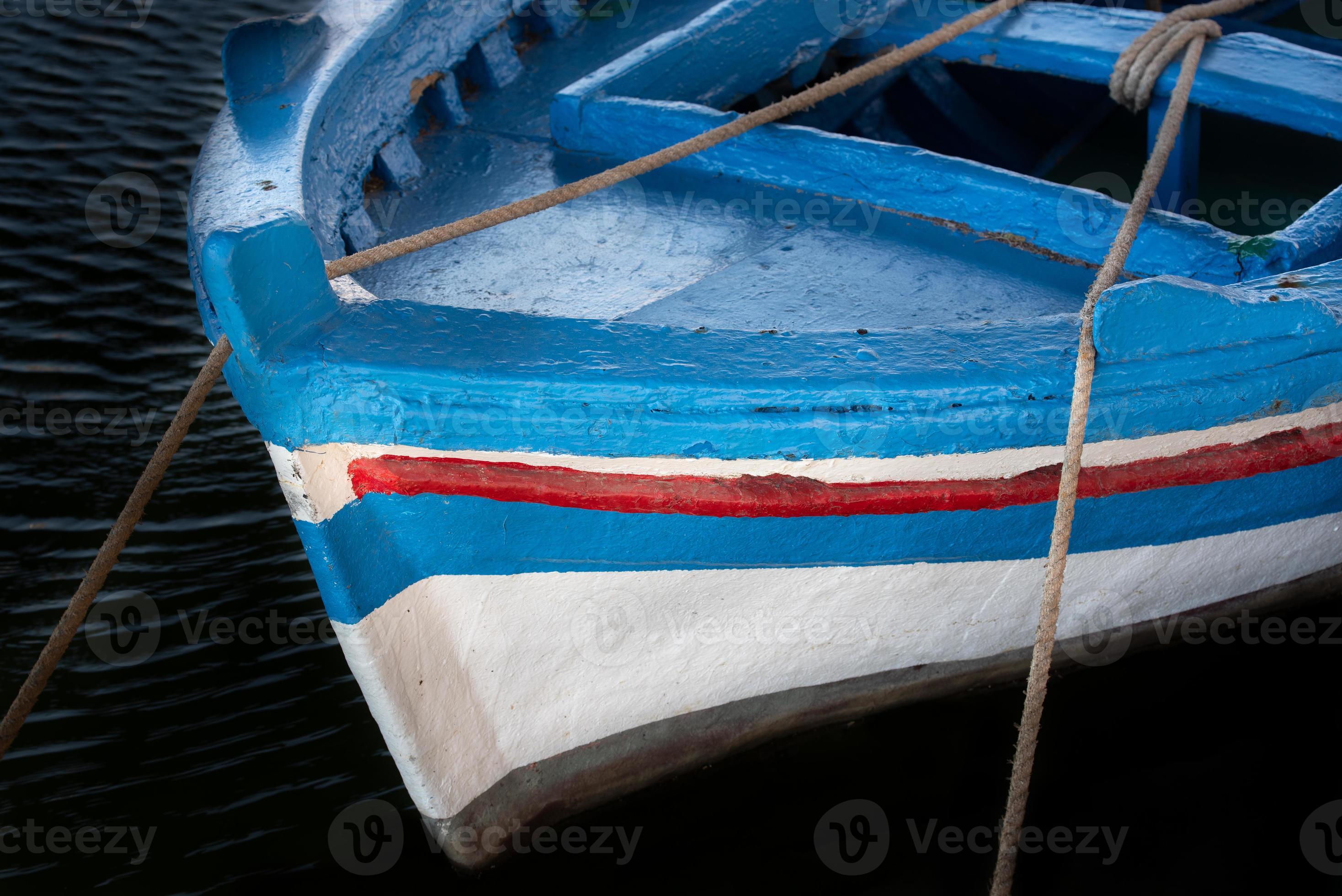 Close up and detail shot of an old wooden fishing boat painted in blue, red  and white paint. The boat stands in the dark water and is tied to the shore  with
