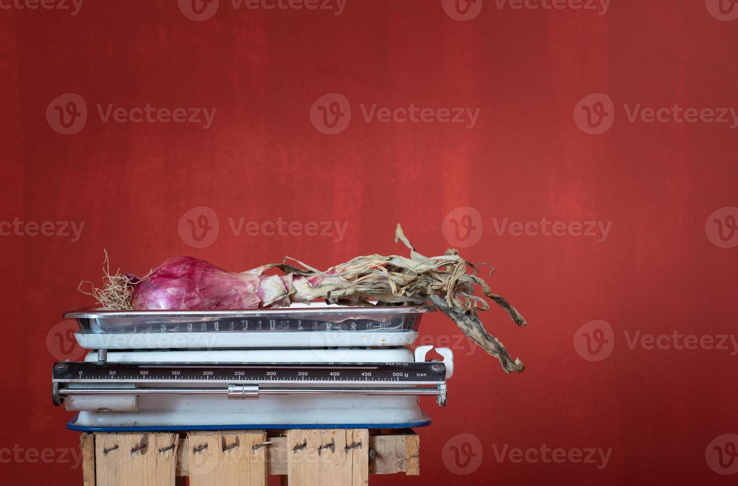 An old kitchen scale stands against a red background with space for text. There is a red onion on the scales. photo