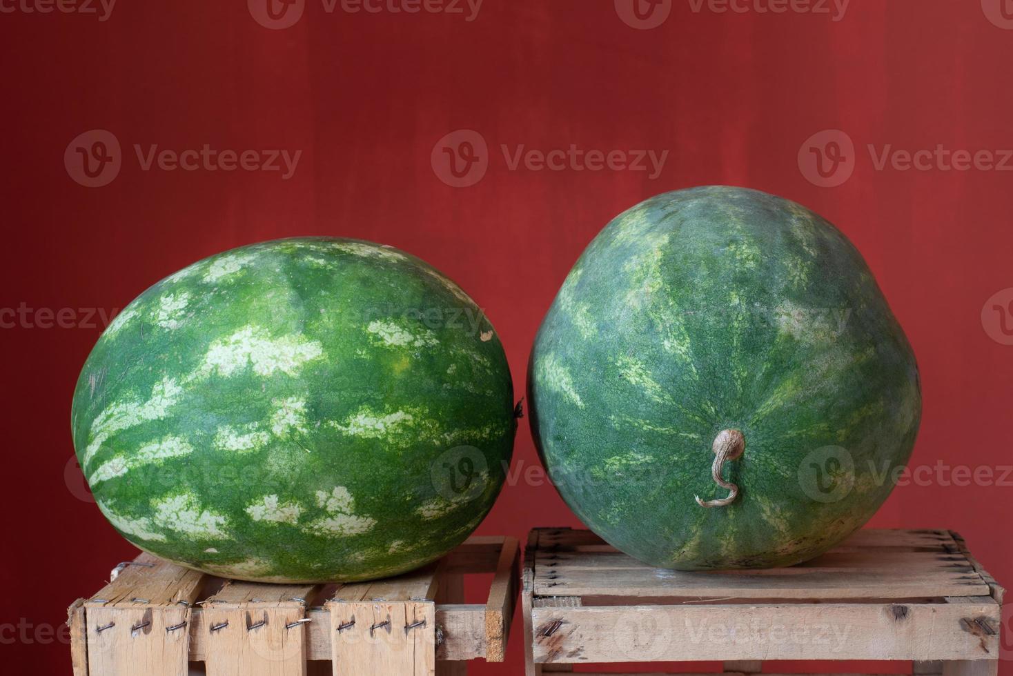 Two green watermelons lie side by side on wooden crates for fruit. The background is red. photo