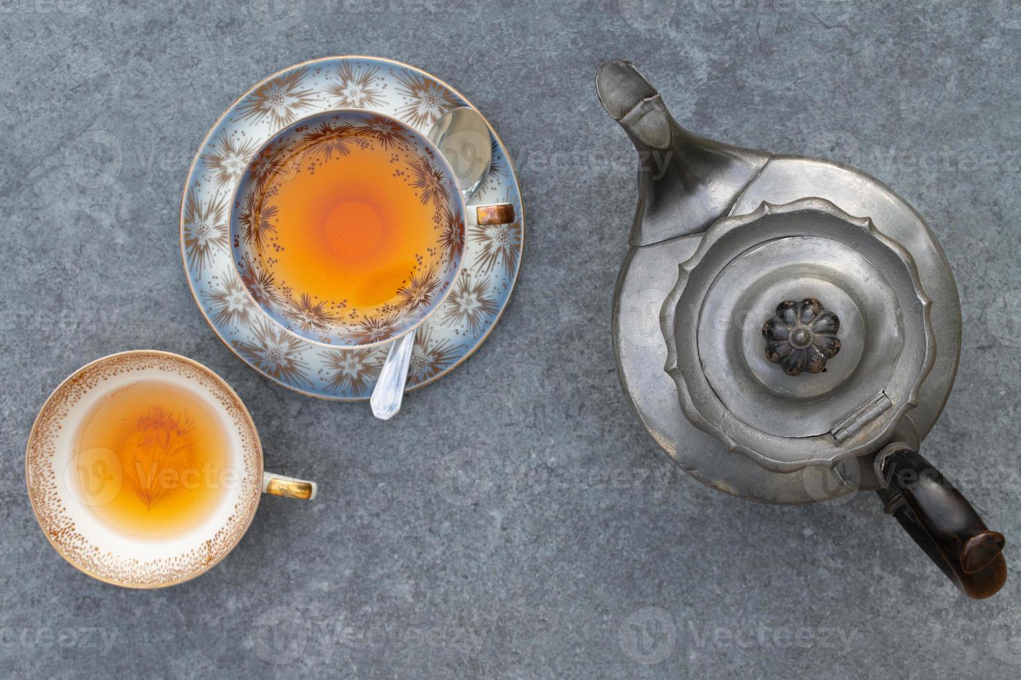 An old metal teapot, an old teacup with tea and saucer and an old teacup without saucer stand next to each other on a gray background. They are photographed from above. photo