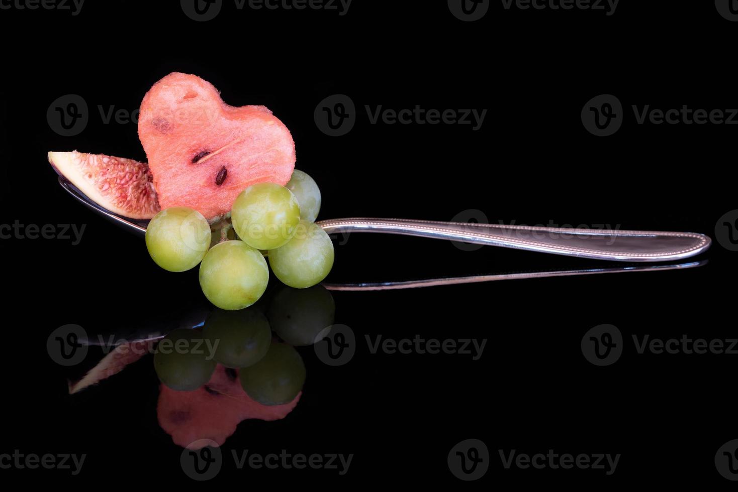 A piece of melon in the shape of a heart, fresh grapes and a piece of fig are placed on a metal spoon. The fruit salad is reflected on the dark background. photo