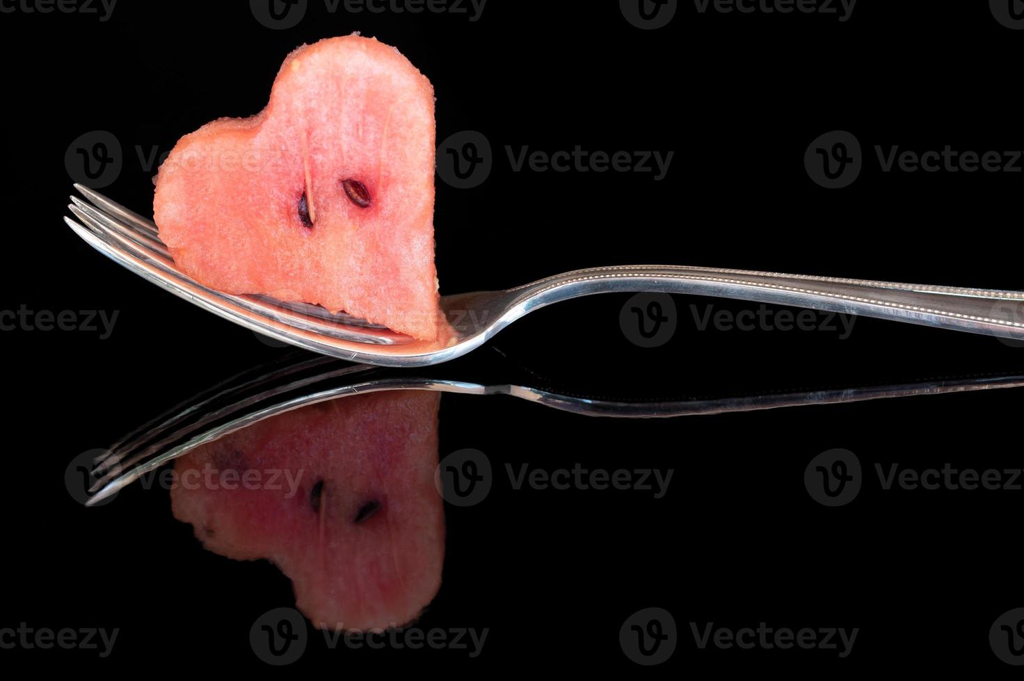 Close-up of a metal fork, with long tines, on which lies a piece of water belone in the shape of a heart. The fork is reflected on the dark background. photo