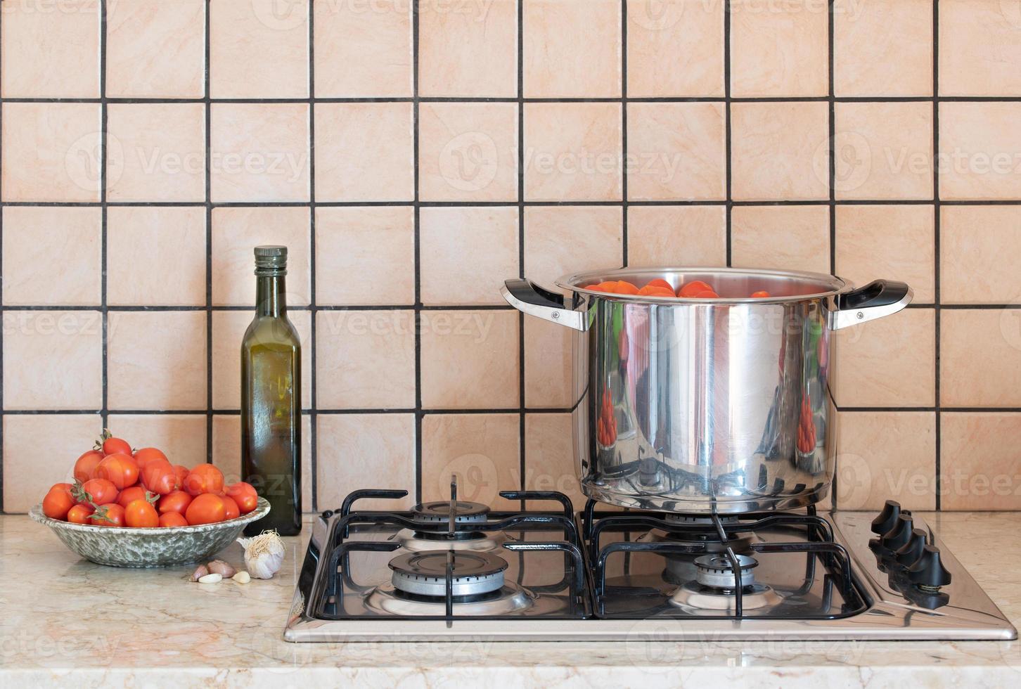 A stainless steel pot with tomatoes in it sits on an old gas stove. Next to the pot is a bowl of tomatoes, a bottle of olive oil and some garlic. The background is tiled. photo
