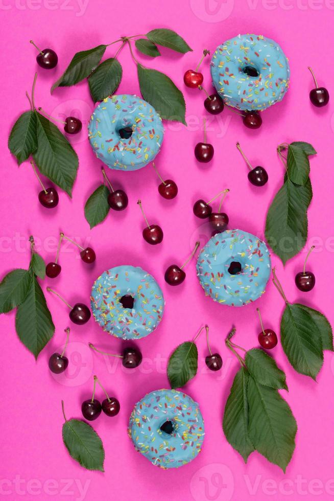 Pink background and still life with crunchy red cherries, cherry leaves and blue sweet donuts lying side by side photo