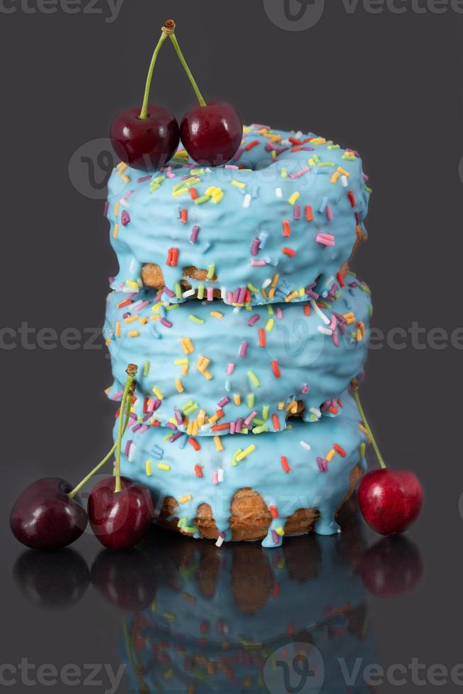 Three colorful donuts with blue icing and love pearls are stacked on top of each other. Above and to the side are red ripe cherries. The background is grey. photo