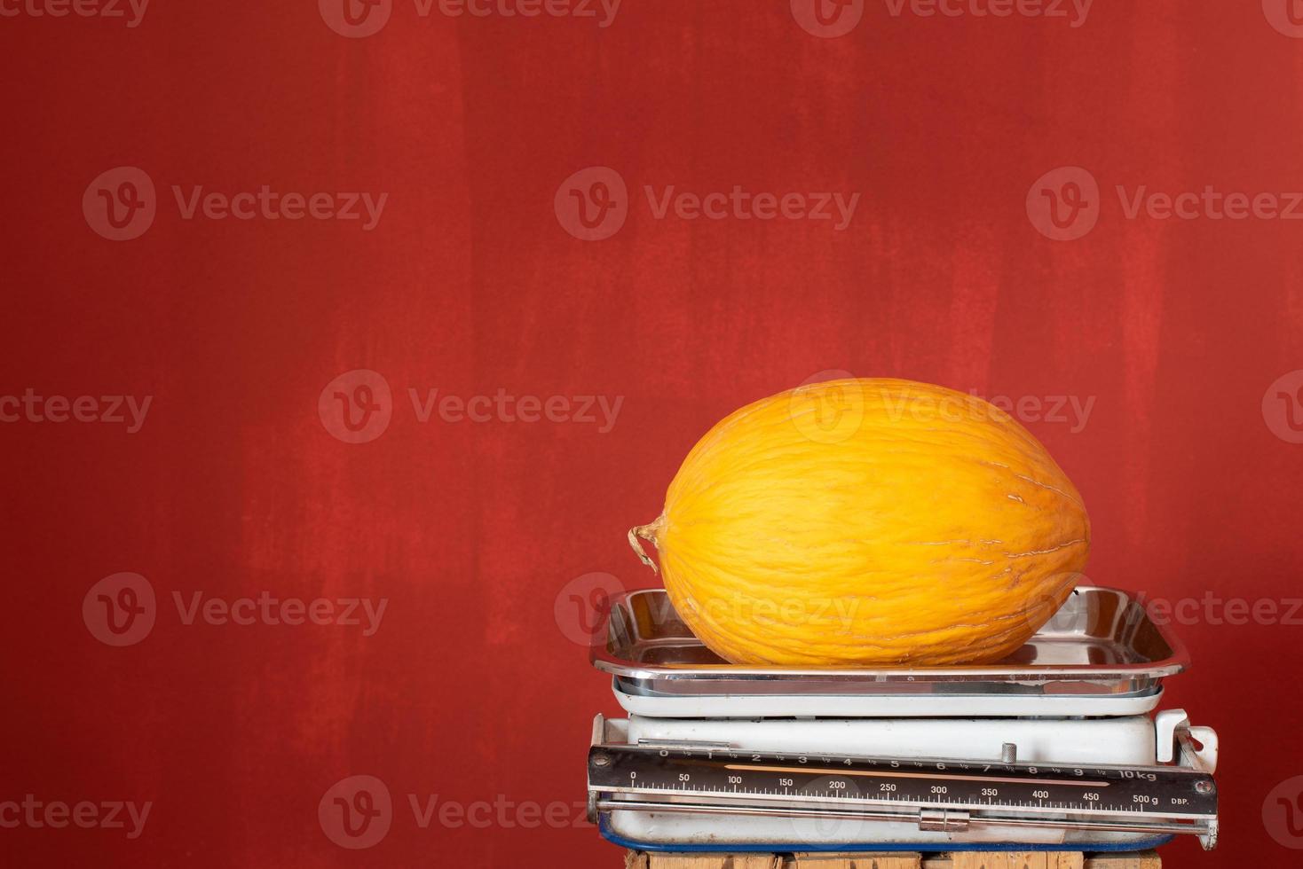 An old kitchen scale stands against a red background with space for text. A yellow honeydew melon lies on the scales photo