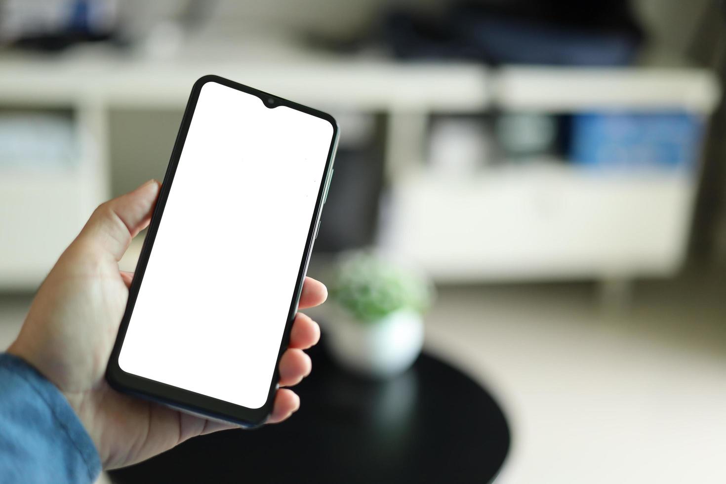 Mockup image of a man holding black mobile phone with blank white screen. In living room at home. photo