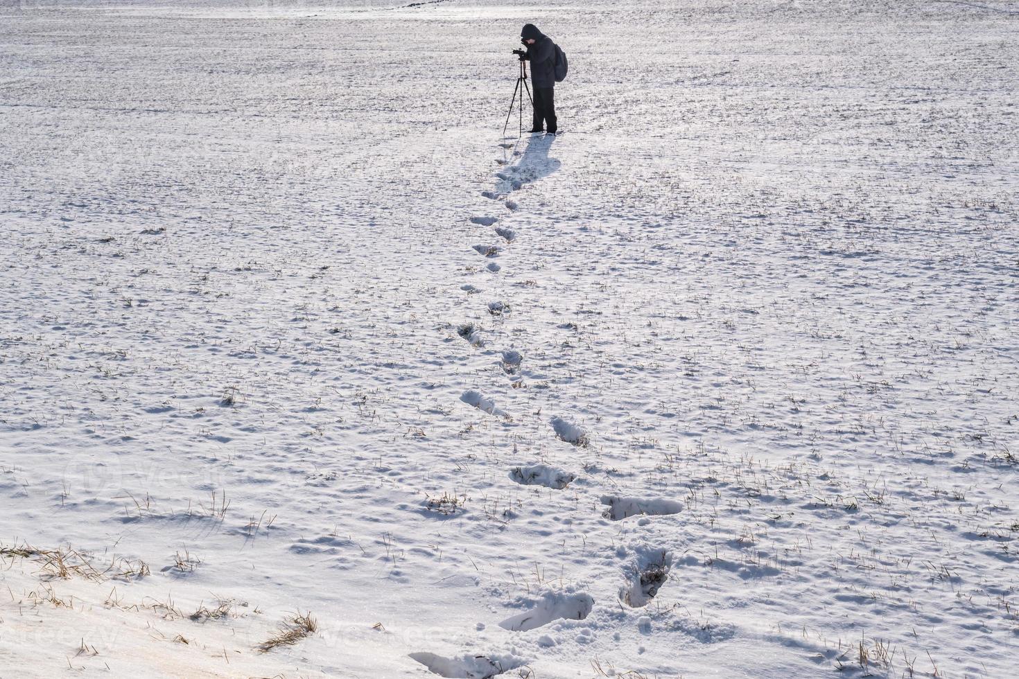 photographer with a tripod in snowy field takes pictures of winter landscape, footprints in snow photo