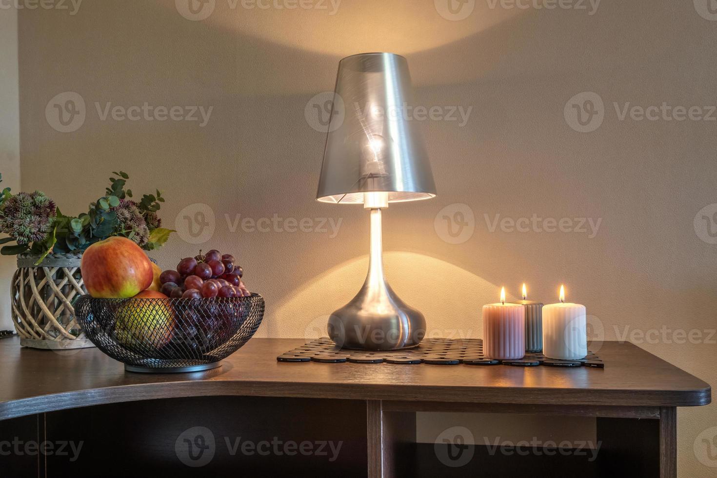 burning candles and a vase with fruit apples and grapes on the table near the lamp photo