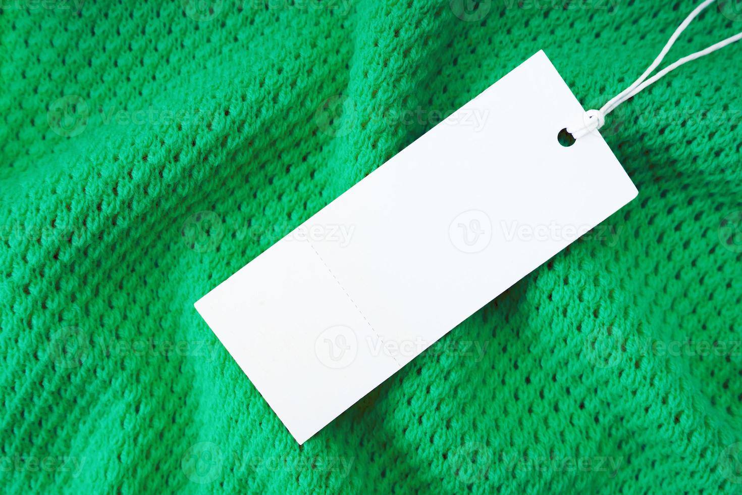 White blank rectangular clothing tag on green knitted fabric background. Shopping, sale, discount mockup photo