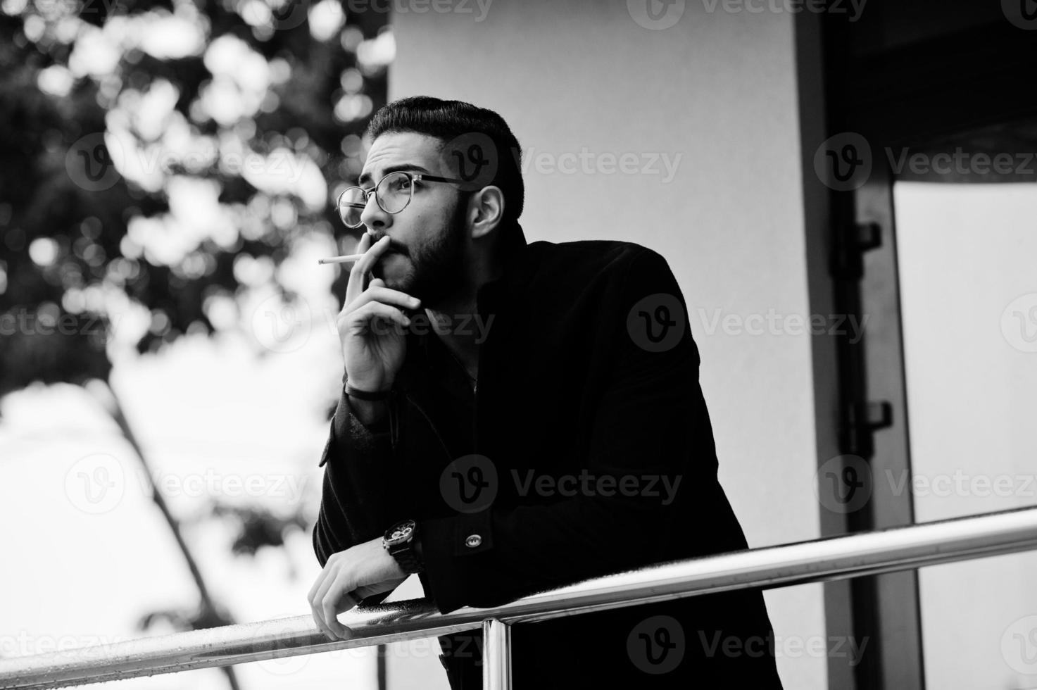 Middle eastern entrepreneur wear black coat and blue shirt, eyeglasses against office building smoking cigarette. photo