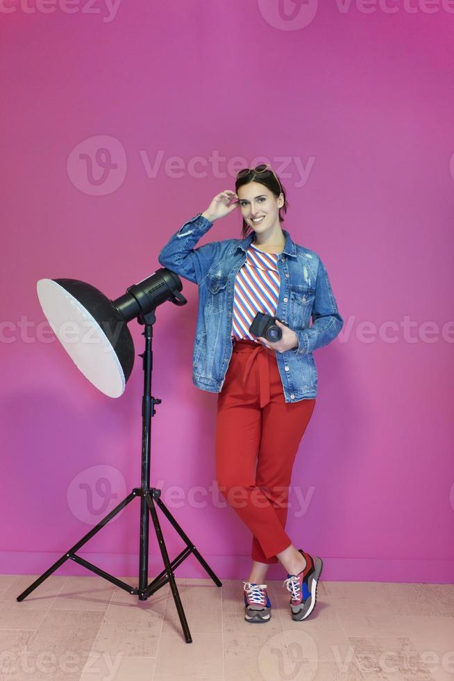 Young woman leaning against studio flashlight and holding camera over a pink background photo