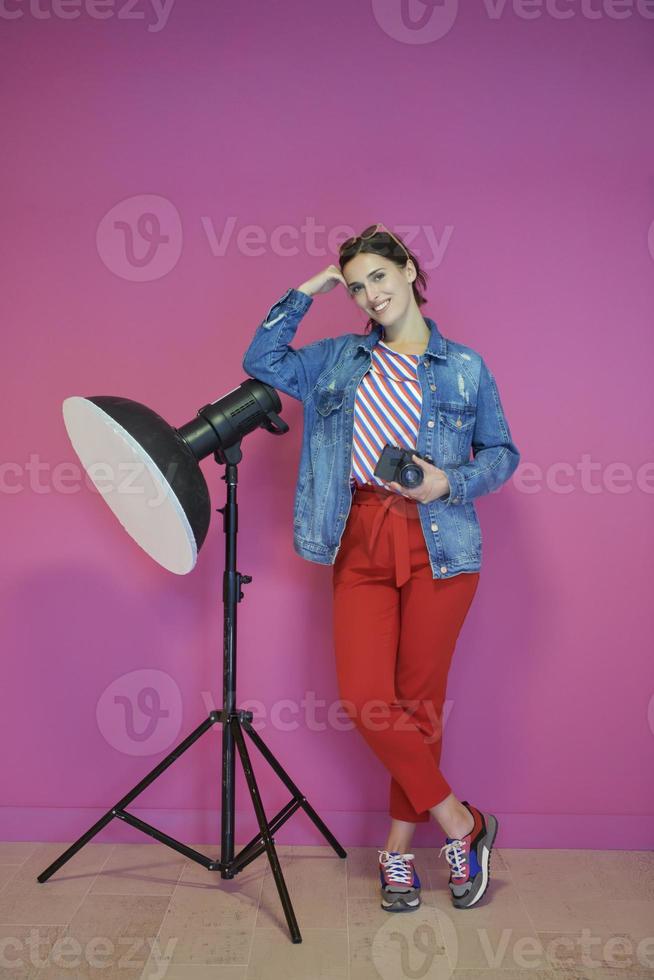 Young woman leaning against studio flashlight and holding camera over a pink background photo