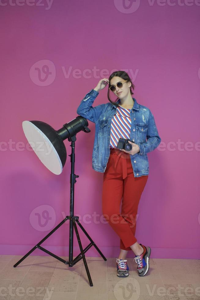 Young woman leaning against studio flashlight and holding camera over a pink background photo