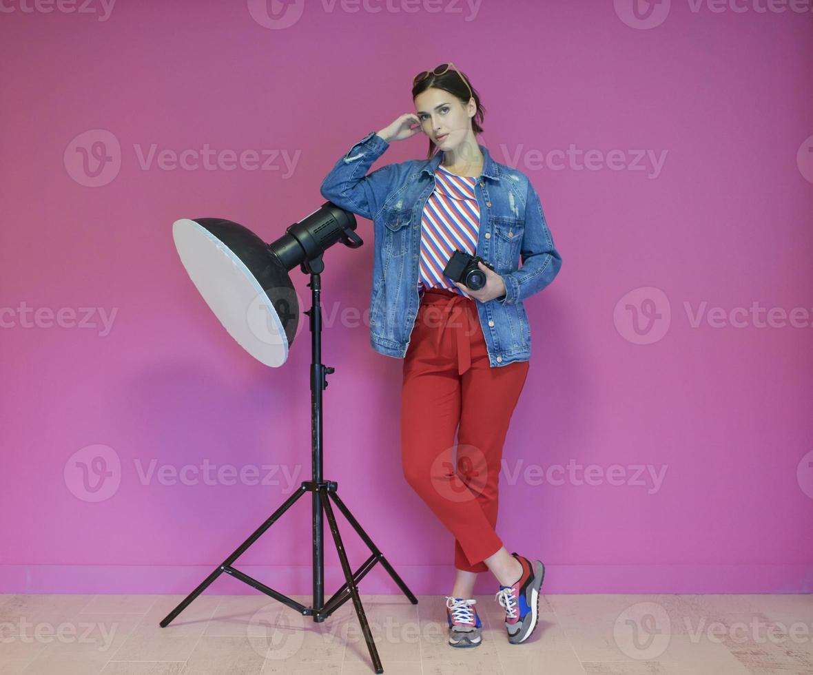 Young woman leaning against studio flashlight and holding camera over a pink background photo