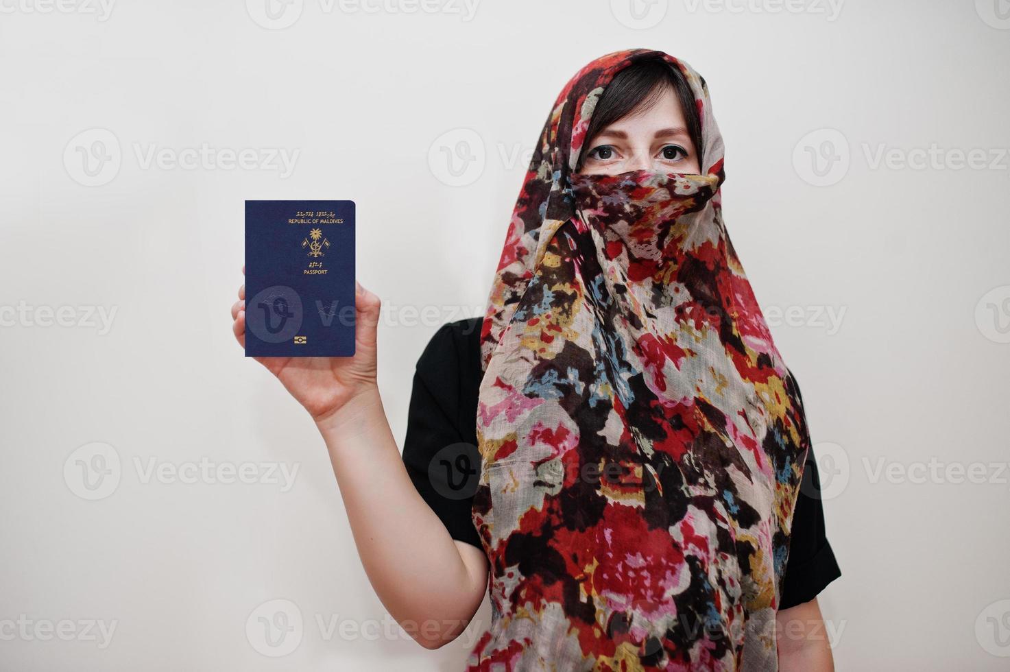 joven musulmana árabe vestida con hiyab tiene pasaporte de la república de maldivas sobre fondo de pared blanca, retrato de estudio. foto