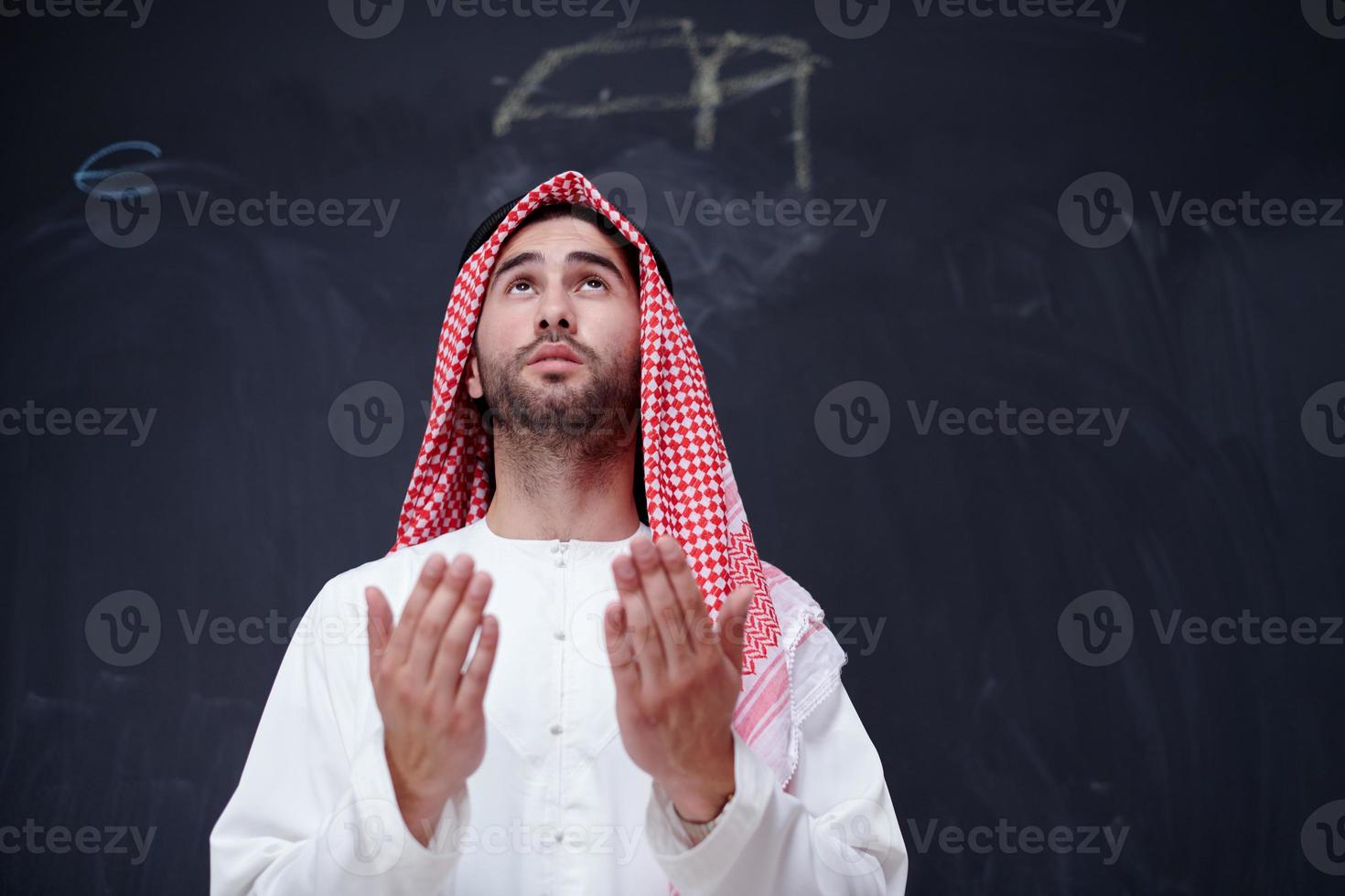 arabian man making traditional prayer to God, keeps hands in praying gesture in front of black chalkboard photo