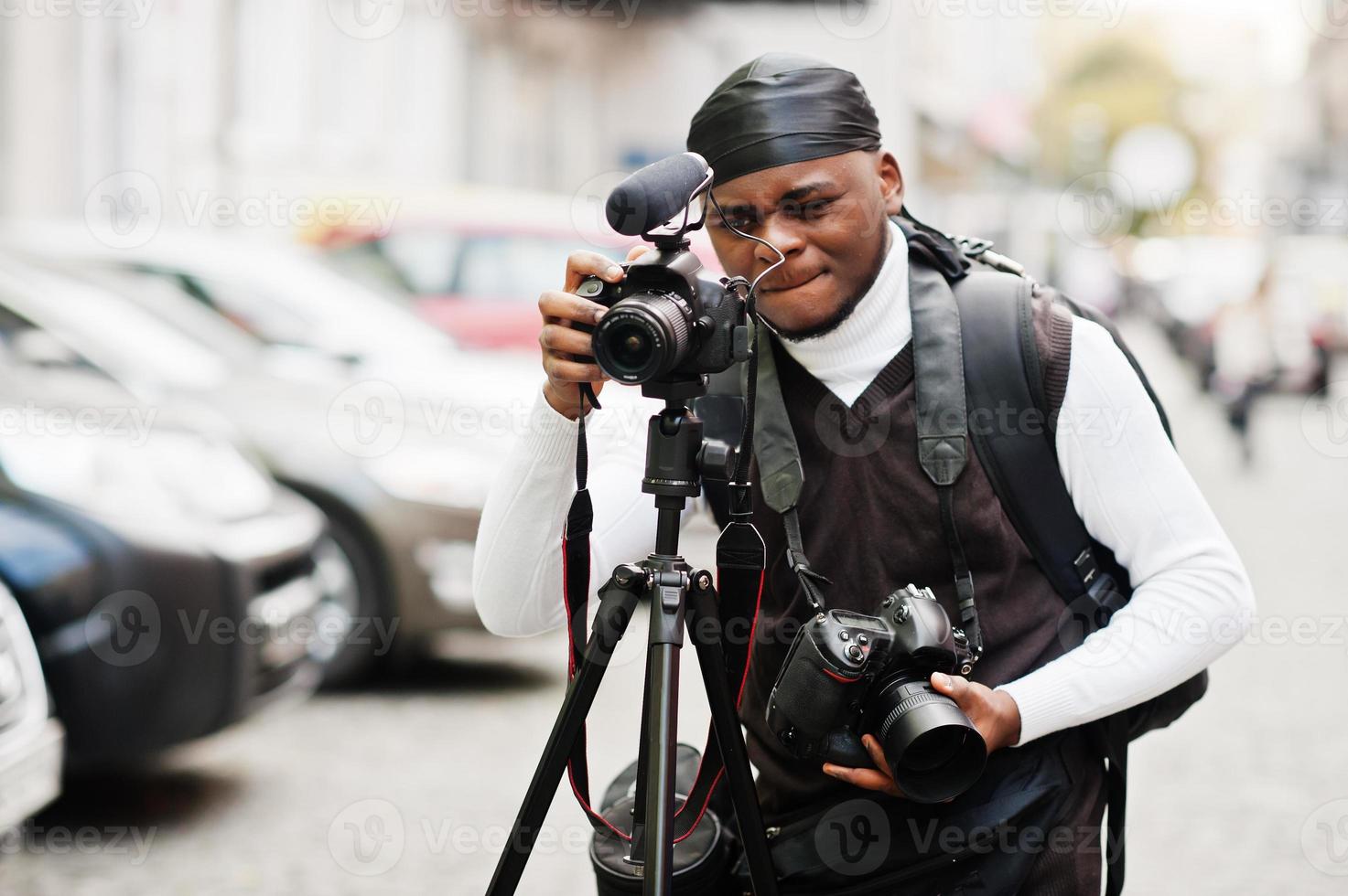 Young professional african american videographer holding professional camera with tripod pro equipment. Afro cameraman wearing black duraq making a videos. photo